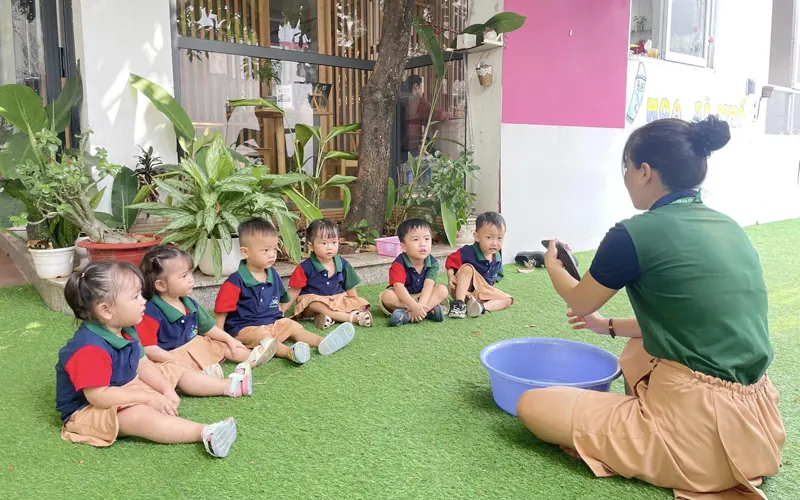 A preschool teacher sitting on a low stool at the edge of a block corner, quietly watching three children build a tall tower, notebook resting closed on her knee, soft daylight